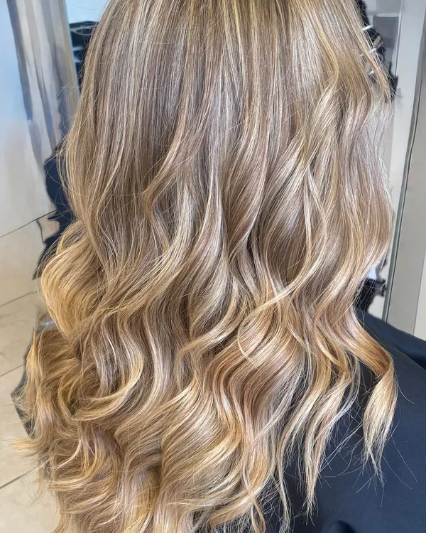 Back view of mid-length hair styled in soft waves, with cool blonde balayage highlights blended through a darker blonde base.